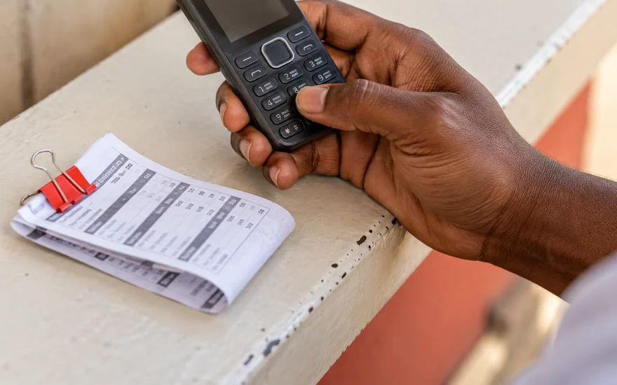 A hand using the physical keypad of a feature phone beside a folded sports slip